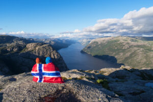 Preikestolen pathway. Couple with the flag of Norway looks at the panorama of the Lysefjord. Tourist attraction. Sunny weather in the mountains