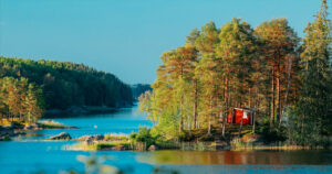 Sweden. Amazing Red Swedish Wooden Log Cabin House On Rocky Island Coast In Summer Sunny Evening. Lake Or River Landscape. Bold Colors.