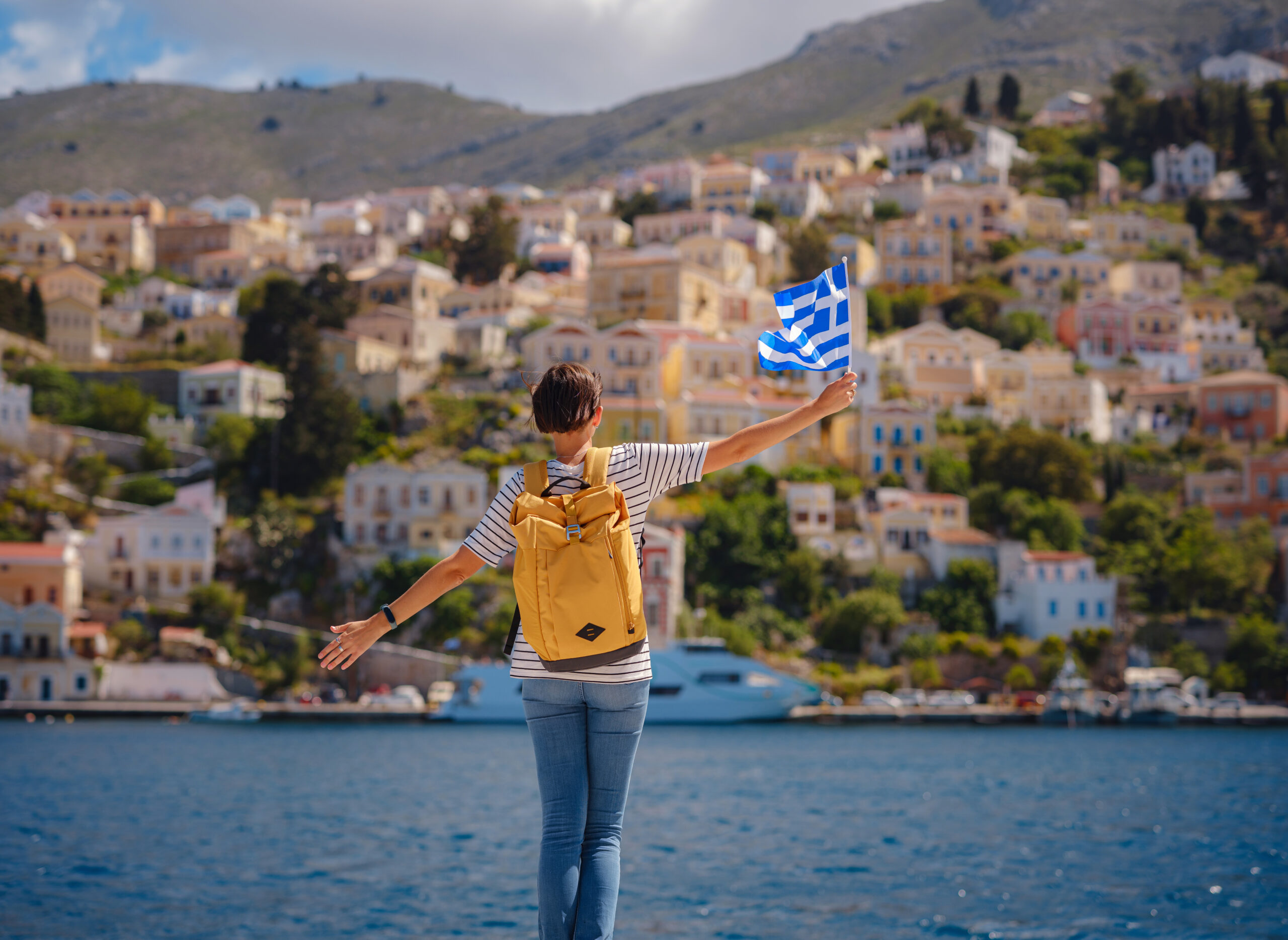 Nice asian Happy Female with backpack anf greek flag Enjoying her holidays on Symi Islands. View of port Symi or Simi, is tiny island of Dodecanese, Greece, calm atmosphere and fabulous architecture.