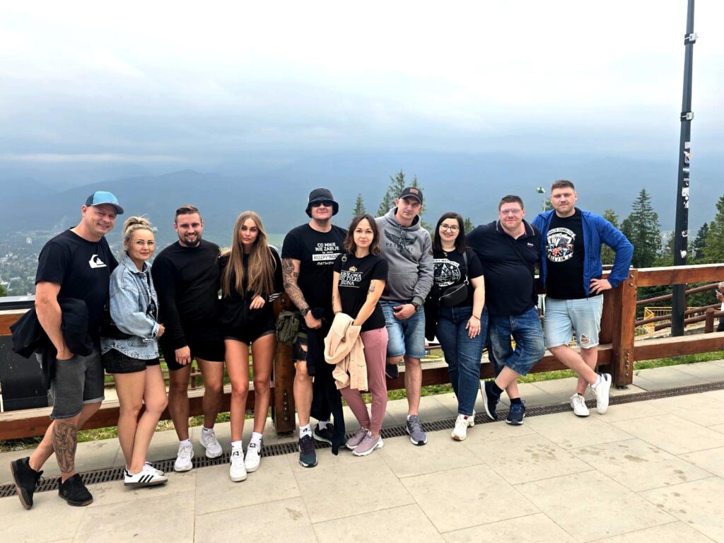 Group of smiling people standing on a scenic mountain terrace with a forested landscape and cloudy sky in the background during a team outing — European relocations team enjoying the view.