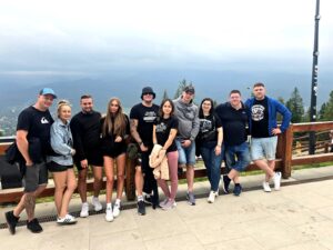 Group of smiling people standing on a scenic mountain terrace with a forested landscape and cloudy sky in the background during a team outing — European relocations team enjoying the view.