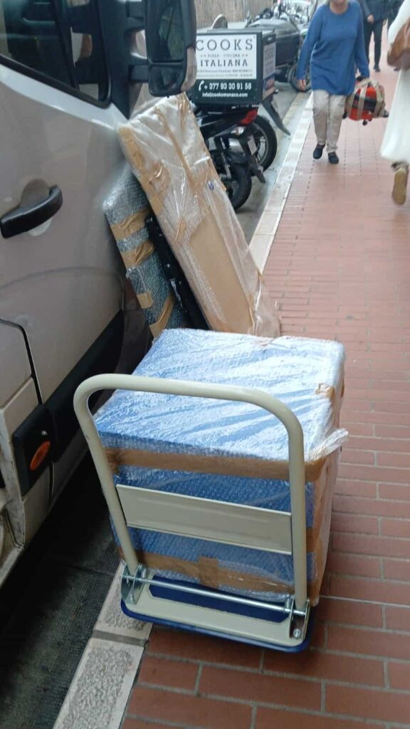 A hand truck loaded with bubble-wrapped boxes leaning against a van on a brick sidewalk, representing cheap removals to Holland.