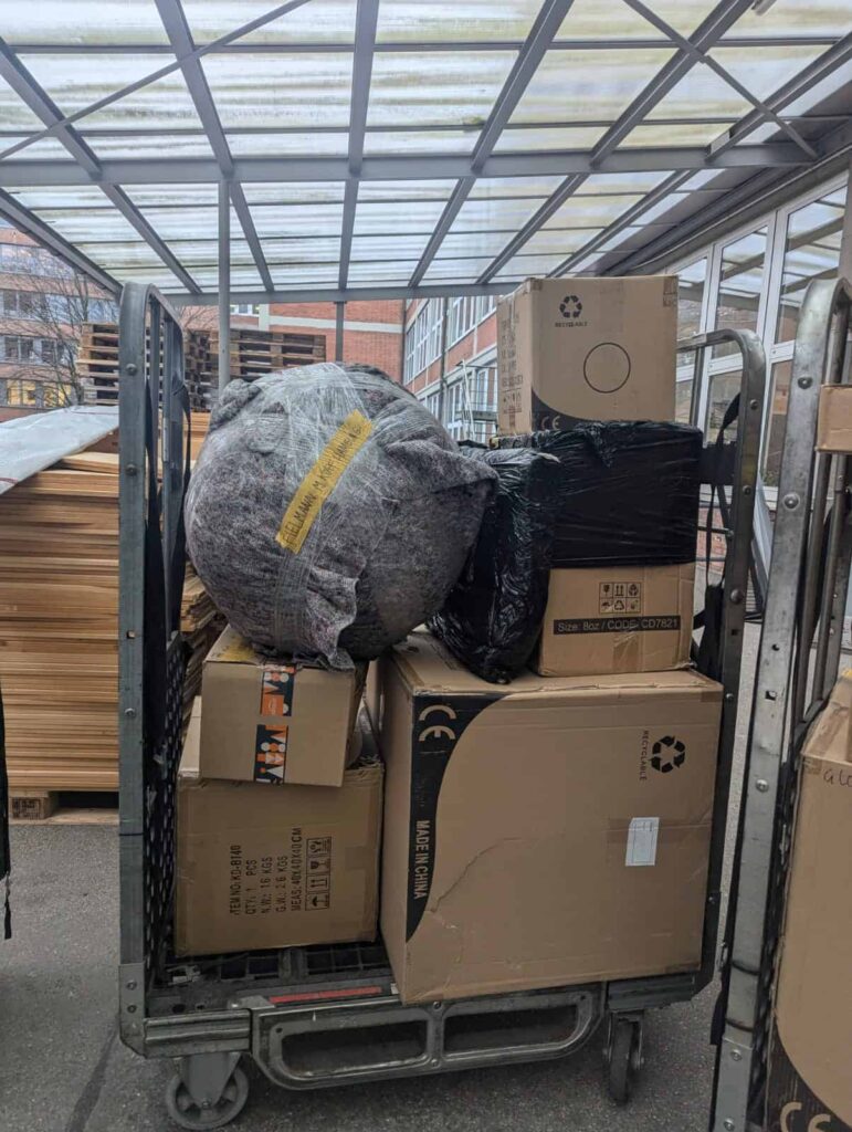 A rolling storage trolley stacked with packed boxes and bubble-wrapped items under a covered loading area, showcasing cheap removals to Holland.