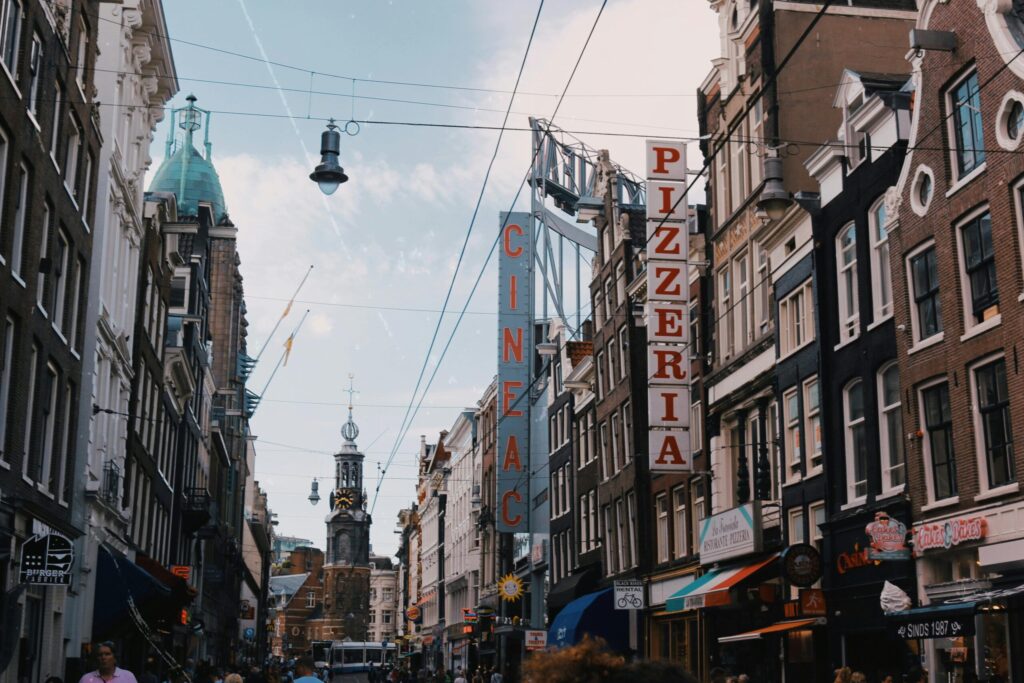 A bustling Dutch city street lined with tall, narrow brick buildings, tram tracks running down the center, and signs for a pizzeria and Cineac cinema; a crowd of pedestrians and cyclists adds to the vibrant scene—perfect for expats in Netherlands looking to immerse themselves in urban life.