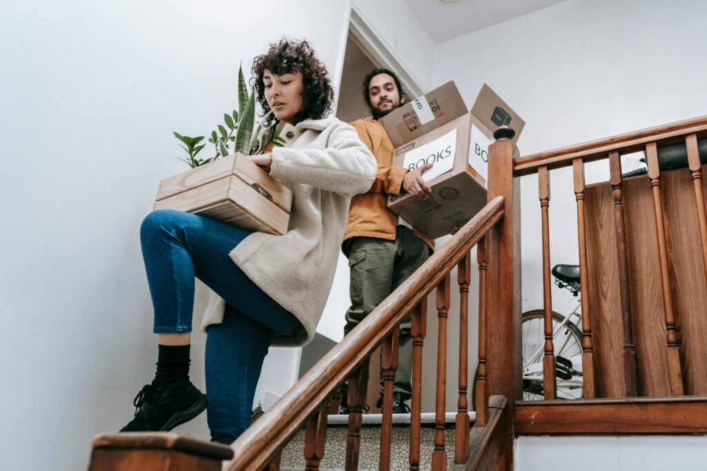 Couple carrying packed boxes and potted plants down a staircase, preparing for their move from the UK to the Netherlands.