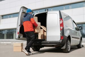 A mover in a blue beanie and red jacket lifts cardboard boxes into the back of a silver van parked outside a modern building, illustrating a relocation to the Netherlands.