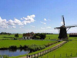 A classic Dutch windmill standing beside a narrow canal, set against a bright blue sky and lush green polder fields with grazing sheep.