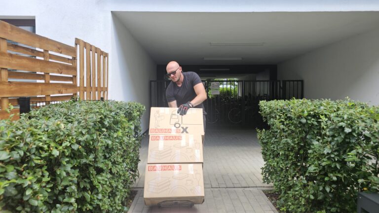 Mover pushing a trolley loaded with large BAUHAUS cardboard boxes along a hedge-lined path toward an apartment building entrance.