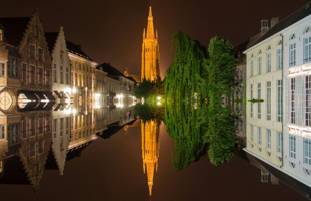 Night view of Bruges canal with illuminated Church of Our Lady and historic Belgian houses reflected in the water, showing the charm of moving to Belgium with VANonsite European removals