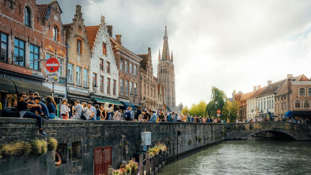 Crowds relaxing by the canal in Bruges, Belgium, with historic brick houses and a stone bridge, showing the lifestyle you can enjoy after moving to Belgium with VANonsite international removals