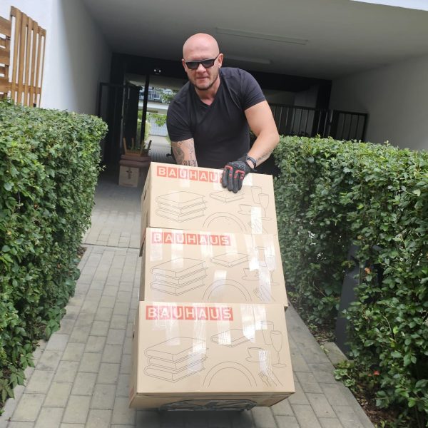 Mover pushing a trolley stacked with three BAUHAUS boxes along a paved path between hedges in front of an apartment building.